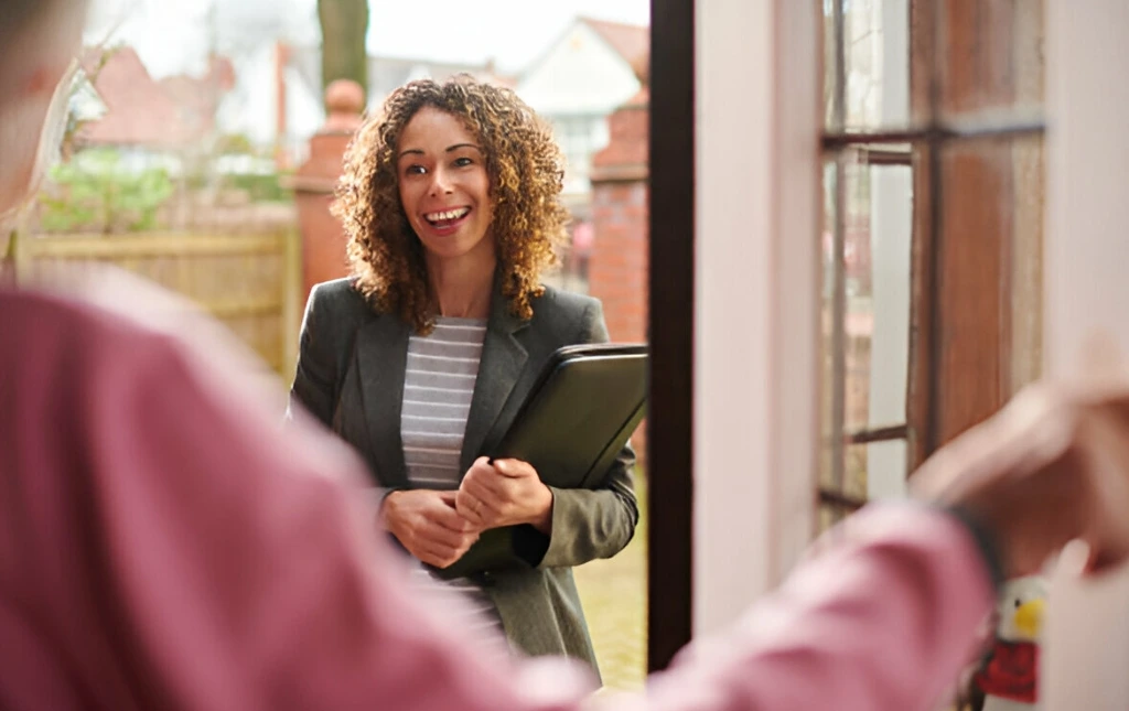 A-professional-woman-is-standing-at-a-door-smiling-and-holding-a-folder-while-greeting-someone-from-inside-the-house