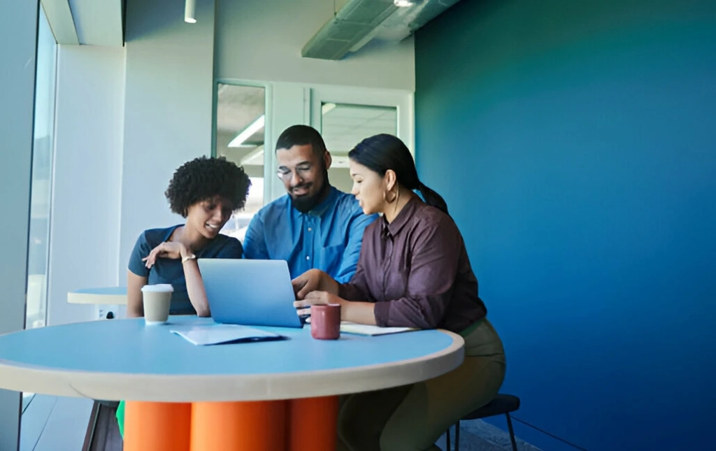 Three-people-are-collaborating-around-a-table-working-on-a-laptop-in-a-modern-office-setting.