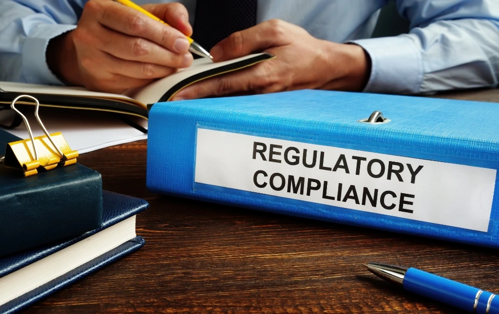 Blue-binder-labeled-Regulatory-Compliance-on-a-desk-with-a-person-writing-in-a-noteboo